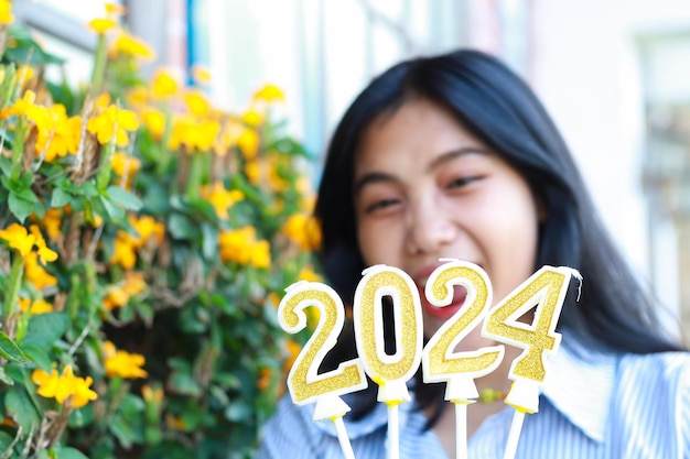 Blurred happy asian young woman show focused 2024 number candle to camera standing beside flower