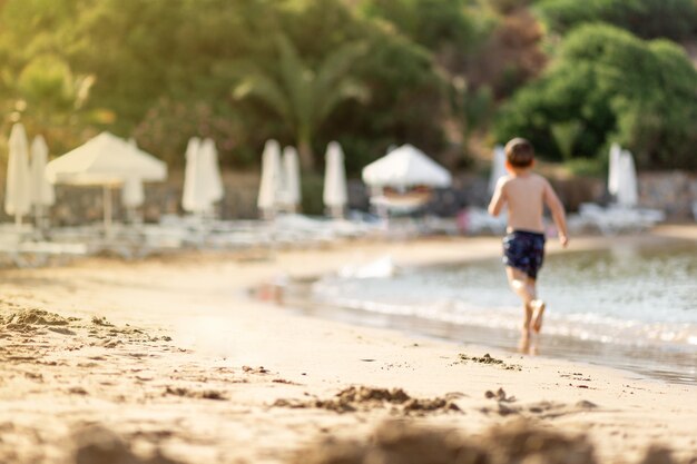 Foto ragazzino sfocato che gioca, corre sulla spiaggia privata vuota durante le vacanze estive. bambini in natura con mare, piante tropicali. bambini felici in vacanza al mare che corrono nell'acqua, isola di cipro