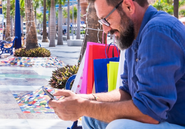 Blurred mature man with black beard resting on a bench after shopping, using mobile phone