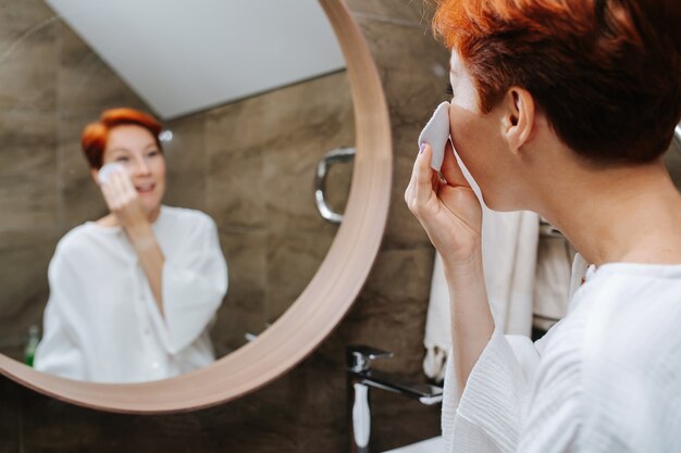 Blurred mirror image of a short-haired mature woman applying\
lotion on her face. she\'s standing in front of the mirror in a\
bathroom, back of her head in foreground.