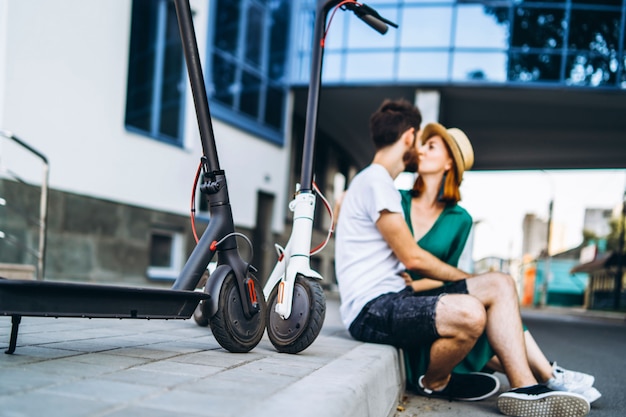 Blurred portrait of man and woman sitting and kissing near a modern glass building. with their electric scooters. In the foreground are their electric