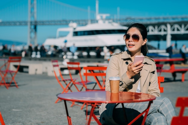 blurred view harbour with yacht in background by oakland bay bridge san francisco under blue sky. beautiful young asian woman in sunglasses reading text message on mobile phone at outdoor coffee shop