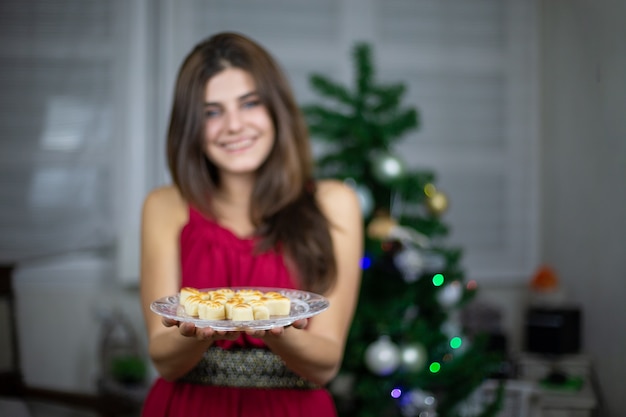 Blurred woman in dress offering Christmas marzipan treat on plate standing indoors