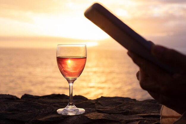 Photo blurred woman's hand standing in front of the sea with a glass of wine looking at her mobile phone while the sun sets in the sea peace solitude relaxation