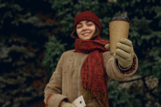 Blurred woman in warm clothes and gloves showing blank paper cup with takeaway coffee