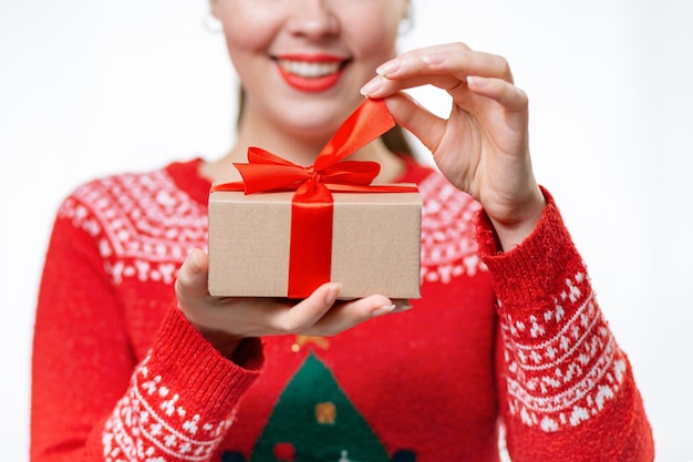 Sfocata una giovane donna sorridente con un maglione di capodanno che tiene un regalo e scioglie un nastro. primo piano delle mani. sfondo bianco. il concetto di shopping natalizio.