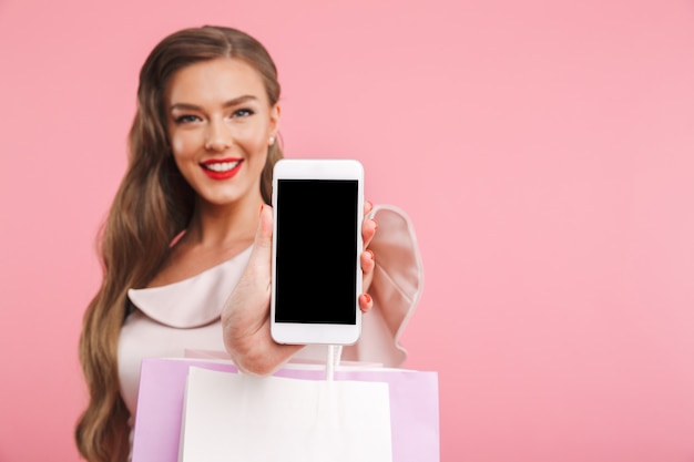 Blurry image of adorable brunette woman smiling and demonstrating copyspace screen of smartphone while holding shopping bags, isolated over pink wall