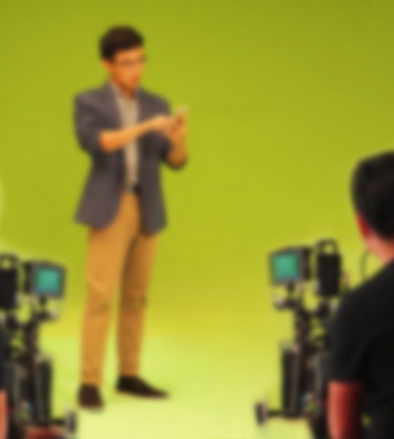 大きな緑色の画面の背景でテレビコマーシャル映画のビデオを作成するぼやけた画像。俳優と一緒に働く映画の乗組員チーム。プロのデジタルカメラと照明セットによる録音。舞台裏の映画