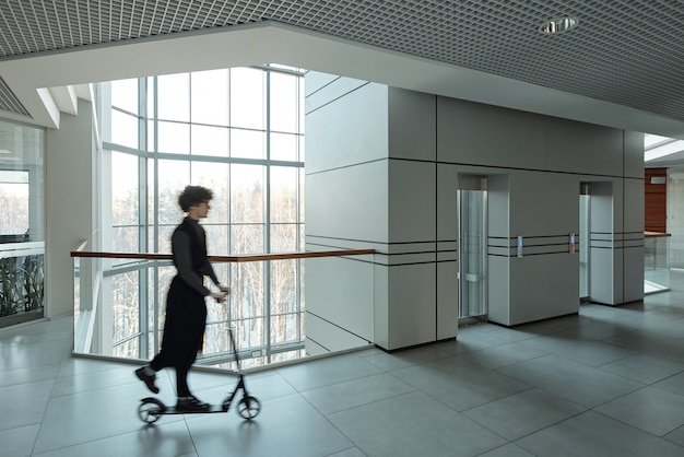 Blurry outline of young active businesswoman riding scooter while moving along corridor inside large office center at break