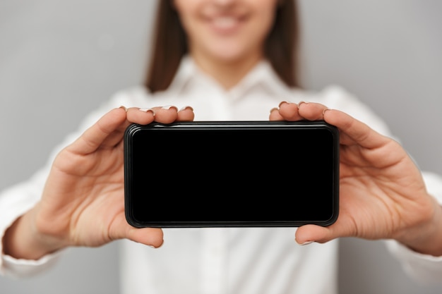 Foto sfocata di donna sorridente con lunghi capelli castani tenendo il telefono cellulare e pubblicità schermo copyspace nero nel primo piano del fuoco, isolato sopra il muro grigio