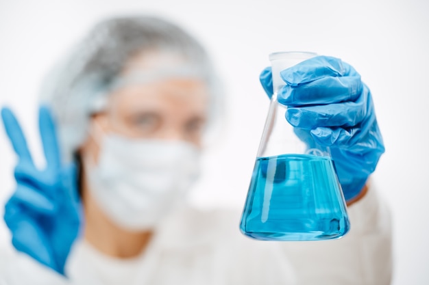 Blurry woman doctor holding flask with blue liquid for coronavirus test shows peace gesture