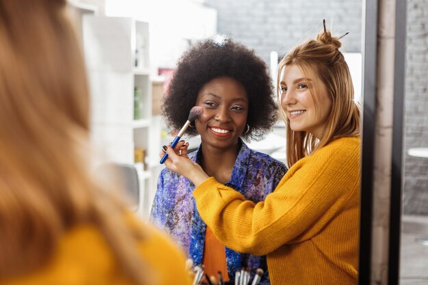 Blusher swatch. Fair-haired smiling stylist wearing a yellow sweater trying on a new blusher swatch on the dark-skinned model