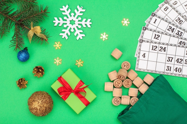 Board game lotto. Wooden lotto barrels with bag and game cards for a game in lotto, Christmas fir tree branches, cones, toy ball and gift box on green background. Top view