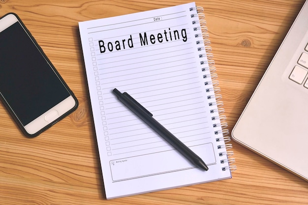 Этикетка заседания совета директоров в блокноте с ноутбуком и смартфоном на деревянном столе. Бизнес-концепция