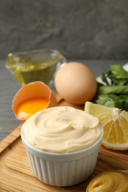 Board with bowl of mayonnaise and ingredients for cooking on wooden background