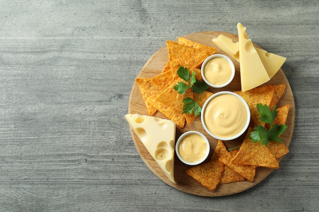 Board with cheese sauce, chips, cheese and parsley on gray textured table