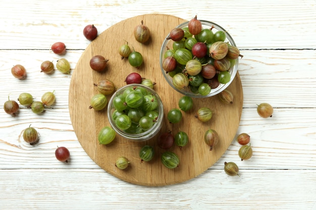 Board with jar and bowl of gooseberry on white wooden background