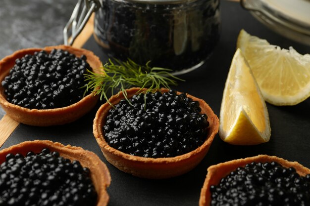 Board with tartlets and jar with caviar on black smokey background, close up