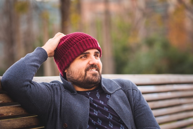 Boarded guy in a woolen hat and sweater sitting on park bench relaxing