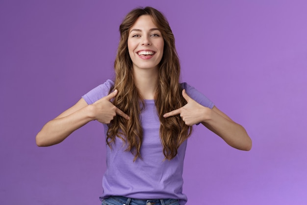 Boastful good-looking curly european female pointing herself proud who she is grinning delighted coming out pride month smiling broadly indicating center chest bragging purple background