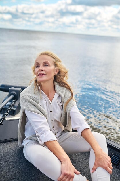 A boat trip. A blonde mature woman in white clothes sitting on a boat and looking dreamy