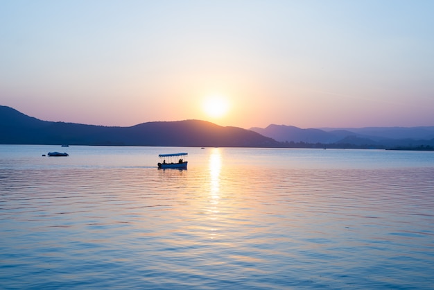 Boats floating on Lake Pichola with colorful sunset reflated on water beyong the hills. Udaipur, Rajasthan, India.