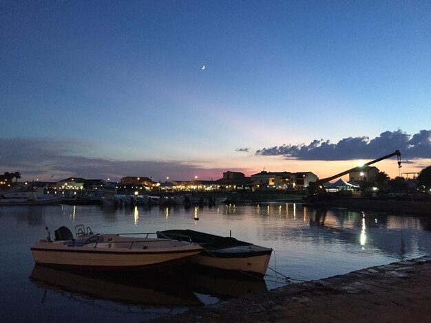 Boats moored in illuminated city against blue sky