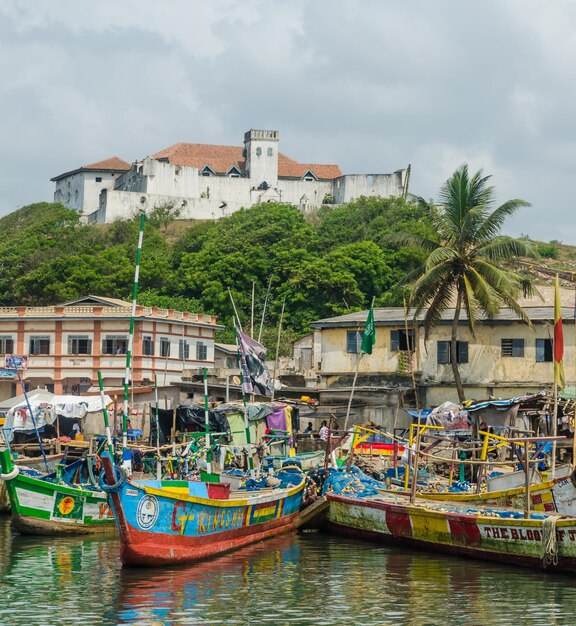 Photo boats moored in water against sky with fort coenraadsburg of elmina in background