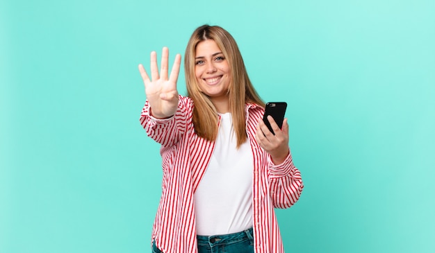 Bochtige, mooie blonde vrouw die lacht en er vriendelijk uitziet, nummer vier toont en een smartphone vasthoudt