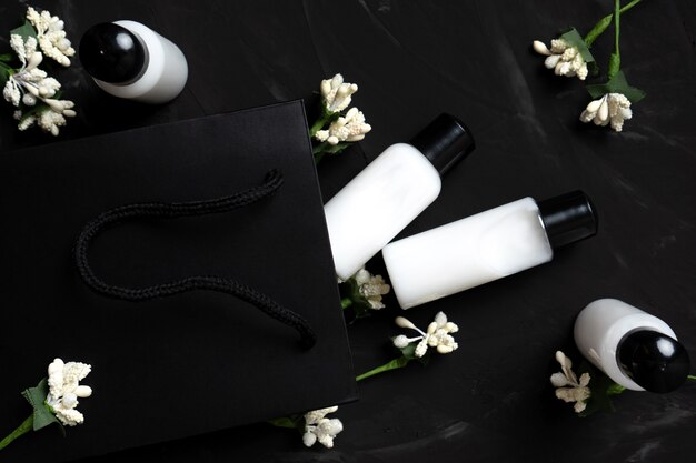 Body care cosmetics in small jars on dark background with paper bag and white flowers
