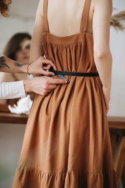 Photo body image of a woman in brown dress, and hands helping to tighten her waist belt. no head. blurred face reflection in the mirror.