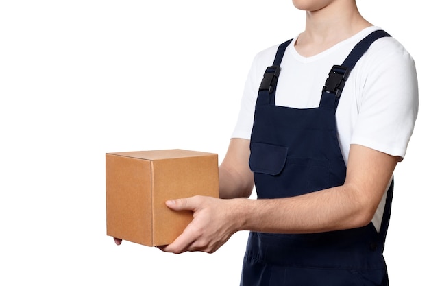 Body of man holding out a cardboard box in front of him and hands it over, isolated on white background. Man with parcel dressed in dark blue overalls and and a white T-shirt. Delivery concept.