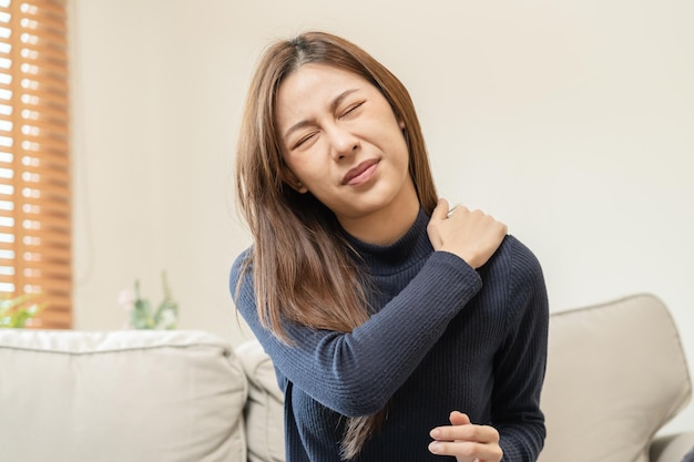 Muscoli del corpo problema rigido asiatico giovane donna attraente ragazza dolore con mal di schiena mal di schiena dal lavoro tenendo massaggiare sfregamento spalla ferita o dolorante seduta sul divano a casa persone sanitarie