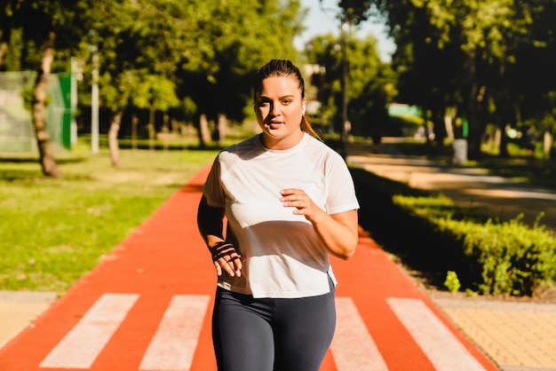 체력 긍정적 인 플러스 크기 한 여성 운동 선수 운동복을 입고 조을 하고 경기장 야외에서 음악을 들으면서 체중을 줄입니다.