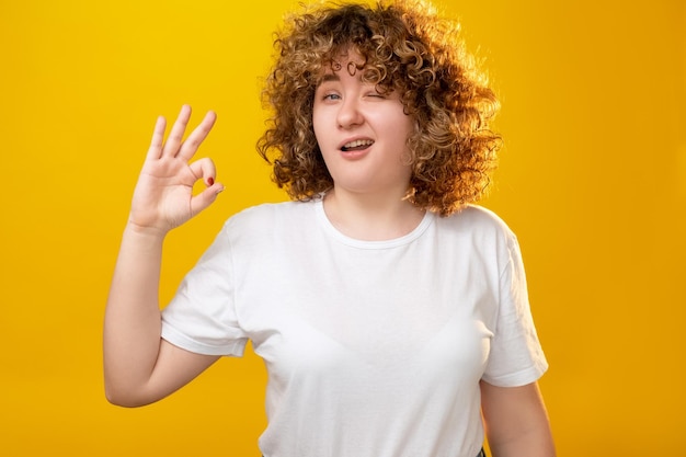 Body positive Supportive gesture Obesity carefree Healthy nutrition Winking joyful overweight young woman with curly hair okay hand sign isolated on orange background