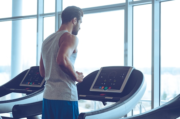 Body power. Young man in sportswear running on treadmill in front of window at gym