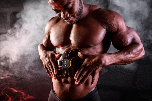 ボディービルダーの男性アフリカ人は、煙のある黒い背景に電子ベルト筋肉刺激トレーナーの腹部の筋肉を使用しています