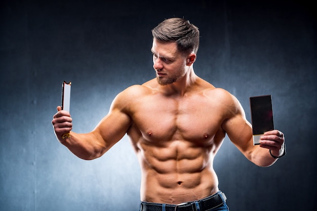 Bodybuilder sportsman holding sport bar and chocolate . Choosing between healthy and harmful food. Naked torso. Grey background. Closeup