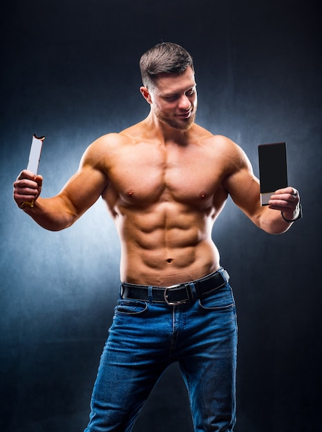 Bodybuilder sportsman holding sport bar and chocolate . Choosing between healthy and harmful food. Naked torso. Grey background. Vertical photo. Closeup