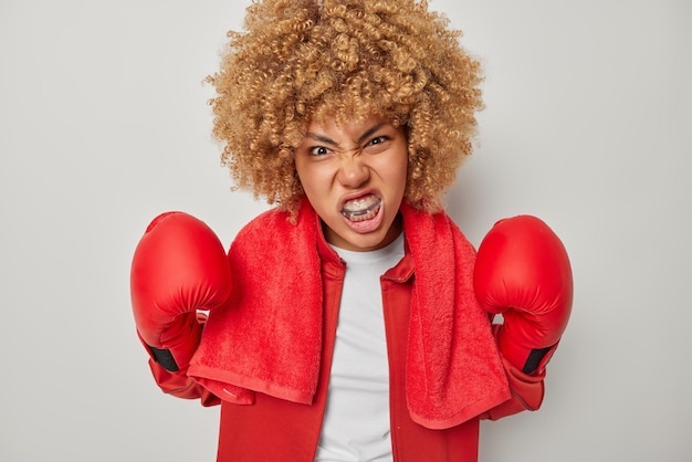 Bodybuilding and sport concept angry curly female boxer wears\
boxing gloves puts on mouthguard ready to fight and become winner\
of championship going to give punch isolated over grey\
background