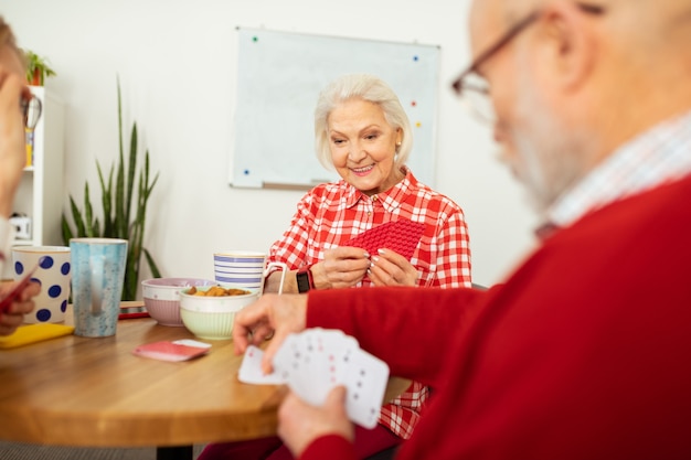 Boeiend amusement. Aangename senior vrouw die haar kaarten vasthoudt terwijl ze met haar vriend speelt