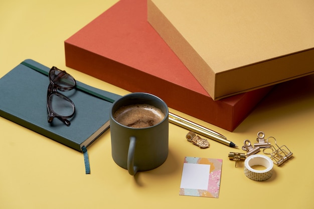 Boek, koffiekopje, leesbril, pen en potloden op geel.