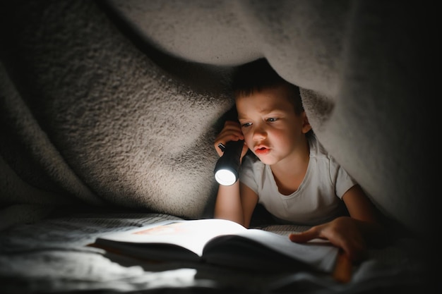 Boek lezen en zaklamp gebruiken. Jonge jongen in vrijetijdskleding die 's avonds in de buurt van de tent ligt.