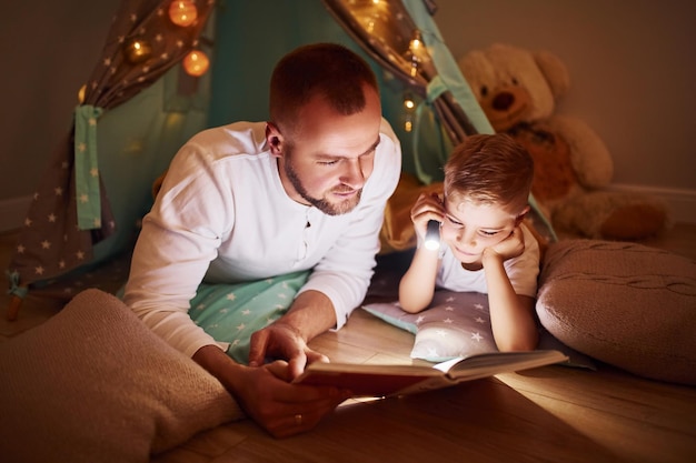 Boek lezen en zaklamp gebruiken Vader en zijn jonge jongen brengen samen tijd door in de buurt van tent