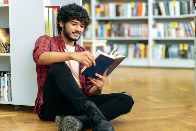 Boek lezen Knappe slimme Indiase of Arabische universiteit of hogeschool mannelijke student zittend op een vloer in een bibliotheek in de buurt van boekenplank lezen interessant boek voorbereiden op examen kennis opdoen glimlach