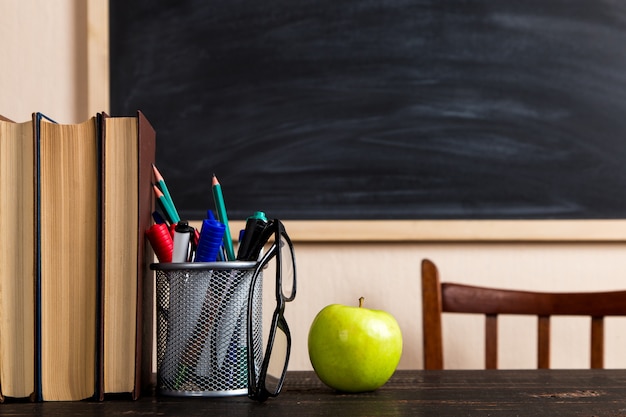 Boeken, appel, pennen, potloden en glazen op een houten tafel, tegen een krijtbord.