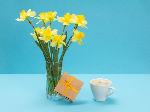 Boeket gele narcissen in glazen vaas een geschenkdoos en een kopje koffie op blauwe achtergrond