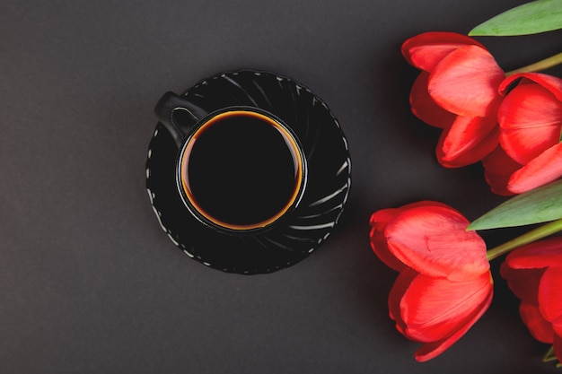 Boeket rode tulpen en kopje koffie op zwart. Plat leggen. Moeder of vrouwendag. Wenskaart. Goedemorgen ontbijt. Kopieer ruimte. De lente.