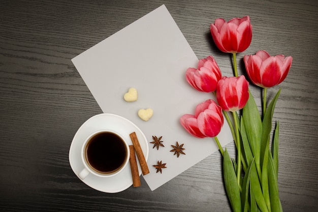 boeket roze tulpen, een kopje koffie, hartvormige suiker, kaneel, steranijs en vel papier op een zwart hout