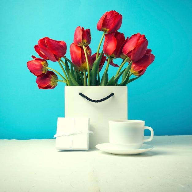 Foto boeket van fel rode tulpen in een witte cadeauzakje, witte kop met koffie, witte geschenkdoos op een blauw. concept gefeliciteerd, verrassingen en geschenken
